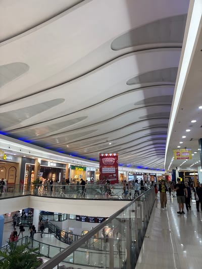 The image shows the interior of a shopping mall in Kelapa Dua, Indonesia. The architectural design is modern, featuring a high, white, wave-patterned ceiling with embedded lighting. Natural light appears to be limited, with the ceiling design likely serving as the primary source of illumination. 

Multiple levels of the mall are visible, each lined with shops and eateries. Glass barriers offer a view of the lower levels from above. People are seen walking along the corridors, visiting shops, and dining in restaurants. Banners advertising "Lunch Deals Voucher" are displayed. Signage for shops and restaurants, including "Anne's", "OLD CHANGKEE", "OMG Chicken", and "MG LIFE", are visible. The photograph suggests a busy and well-maintained commercial environment.
