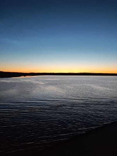 The image depicts a serene lake at dusk, with the sky transitioning from a deep blue at the top to a vibrant orange near the horizon. A dark, silhouetted tree line separates the water from the sky. The water's surface is gently rippled, reflecting the twilight colors. The foreground shows a dark, indistinct edge, possibly a shoreline or a dock. The scene appears to be located in the vicinity of Irdomatka, Russia, given the natural landscape. There are no people or animals visible in the image.