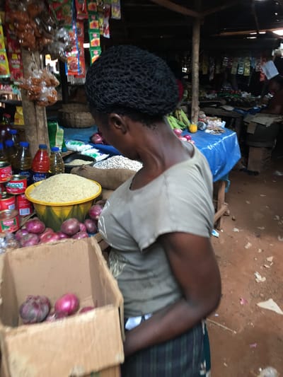 The image provides an angled, overhead view of a market stall in Enyibichiri, Nigeria. The primary visible subject is a person, likely a vendor, with dark skin and dark, tightly curled hair, wearing a light-colored (beige or tan) sleeveless top. The person's head is bowed as they look towards the goods.
The stall is packed with various items. In the foreground and mid-ground, numerous red onions are prominent, some loose and some within a cardboard box. A large yellow bowl filled with white rice is centrally placed, surrounded by other produce like green bananas. Bottles of cooking oil and bottled water, including one clearly labeled "Aquafina," are visible, alongside stacks of red cans, possibly a brand like "Mama's Choice," and other packaged goods.
The background reveals shelves laden with various snacks, candies, and other small consumer items, creating a dense display typical of a local market. The stall structure appears to be made of wood, and a blue tarp is visible, likely serving as a cover or part of the stall's enclosure. The ground is unpaved dirt. The scene depicts a bustling, informal marketplace, likely during daylight hours, showcasing a variety of food and household provisions.
