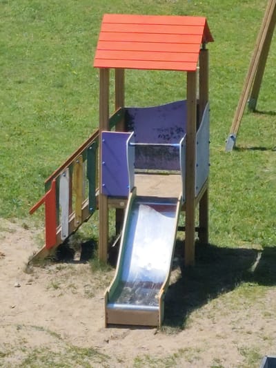 The image shows a children's playground slide with an orange roof. The slide is made of metal and has a wooden frame. There is a set of stairs leading up to the slide. The playground is surrounded by grass and sand. There are no people in the image. The location of this playground is not provided in the image.