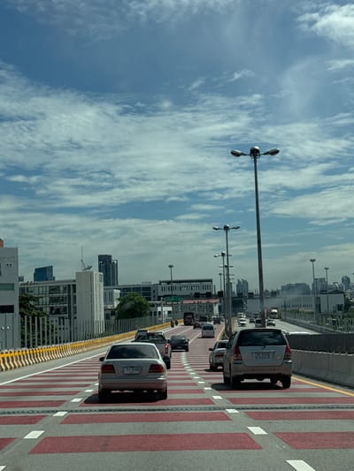 The image depicts a view from a vehicle traveling on a multi-lane highway, likely elevated, under a partly cloudy blue sky. The highway features prominent red and white striped road markings. Numerous cars are visible on the road, including a silver sedan in the foreground with Thai license plate 'ทพ 2256' and a silver minivan in the middle ground with license plate 'MD 8224'. On either side of the highway, there are concrete barriers, streetlights, and buildings, indicating an urban environment. In the distance, several high-rise buildings are visible. The location is identified as Song Khanong Subdistrict, Thailand.