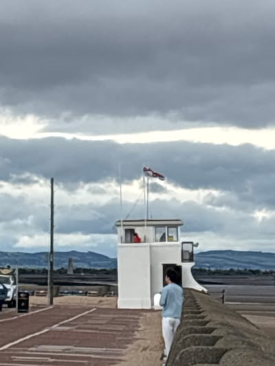 The image depicts a vertical scene featuring a white lifeguard tower against a backdrop of dramatic, cloudy skies. The tower has a small enclosed upper level with a window, through which a person wearing a light blue top and white trousers is visible, leaning forward as if looking down. A flag with red and white markings is attached to a pole in front of the tower. The foreground shows a textured, reddish-brown surface, possibly a pier or boardwalk, with a dark railing on the right side. In the distance, hills or mountains are faintly visible under the overcast sky, suggesting a coastal or lakeside setting. The overall impression is of a possibly breezy or stormy day, given the heavy cloud cover. The scene appears to be set during the daytime, with diffused light. There are no visible text elements in the image.