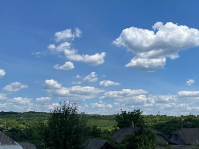 A wide shot captures a vibrant blue sky filled with puffy white clouds, scattered above a verdant landscape. In the foreground, the roofs of several residential buildings are visible, some with weathered, grey shingles. Dense, green trees and lush foliage dominate the middle ground, forming rolling hills that extend into the distance. The horizon is dotted with additional trees and what appear to be utility poles. The scene suggests a rural or semi-rural setting, likely within the city of Lipcani, Moldova, on a clear day. No people or animals are present in the frame.