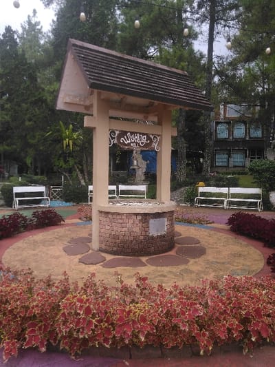This image captures an outdoor park-like scene featuring a wishing well structure in Ede, Netherlands. The central object is a beige-colored wishing well with a dark brown shingled gabled roof supported by four vertical posts. Suspended from the front beam of the structure is a decorative wooden sign bearing the stylized text "Wishing Well," with a small, dark-colored bell hanging directly below it. The well's base is cylindrical, constructed from reddish-brown bricks, and topped with a grey coping stone. A small, square, light-colored plaque is affixed to the front of the brickwork.
The wishing well stands on a raised, circular, tan-colored concrete platform, around the base of which are four irregularly shaped, dark brown stepping stones. This platform is encircled by a bed of low-lying plants exhibiting variegated red and light green foliage. Beyond this, the ground features a multi-colored, segmented pattern in shades of purple, blue, and yellow.
In the mid-ground, three white-painted metal park benches are visible: two to the left and one to the right, positioned behind the well. The background is characterized by numerous tall evergreen trees, primarily pines, under an overcast sky. Several white, pear-shaped decorative light bulbs are strung overhead between the trees. To the right, a two-story building with a dark facade and multiple light blue arched windows on its upper floor, along with rectangular windows on the lower floor, is partially visible. The scene contains no individuals, actions, or overt interactions.