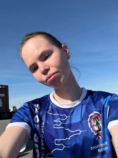 A fair-skinned person is pictured outdoors against a bright blue sky. The person wears a blue t-shirt with a white collar and white trim on the sleeves. The shirt has a pattern of darker blue geometric shapes. On the front of the shirt are white images of building silhouettes that resemble orthodox churches. There is lettering above the silhouette image that reads, "SPORTTOUR RUSSIA". Above that is a logo with a red and white design. The bottom of the shirt is only partially visible and shows reverse lettering, "SID 2026 MY TYP".
The location is Verkh-Sosnova, Russia. Other objects in the scene include a dark colored building and a black vehicle. A utility pole is visible in the distance.