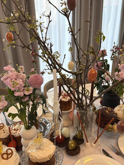 A photograph depicts a festive table setting, likely for Easter, indoors. There are no people visible. The focal point is a centerpiece of bare branches decorated with colorful eggs. Pink flowers are also part of the arrangement. The table is covered with a white tablecloth and set with plates, silverware, and glassware. Several Easter cakes, some decorated with white frosting and pretzels, are present. A tall, slender candle stands in a silver candlestick. The background includes a window with sheer, gray curtains. The photo appears to be taken in a private home, rather than a public space. Beketovo, Russia is not apparent in the image.