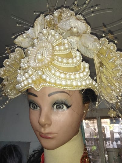 A female mannequin head, displaying an elaborate, multi-layered headdress predominantly in off-white, cream, and pale yellow, is positioned in an indoor setting. The headdress is extensively adorned with numerous faux pearls of varying sizes, arranged in concentric rows, floral motifs, and clusters. Fabric leaf-like and floral elements, some with beaded edges, are integrated into the design. Thin, flexible wire-like structures, each tipped with small pearls, extend upwards and outwards from the headdress. The mannequin's face features gray eyes with heavy black eyeliner, white inner liner, and painted black eyelashes. Its eyebrows are dark and arched, and its lips are pale pink. A light cream-colored collar covers its neck, below which a segment of a red and gold beaded necklace or garment is visible. The background to the left is dark and indistinct. To the right, a multi-paned window reveals blurred outdoor structures and vegetation, suggesting an urban or semi-urban environment. The image depicts a static display. The location is specified as Laguna, Costa Rica.