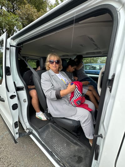 The image shows the interior of a white van with its side door open, revealing several people seated inside. In the foreground, a woman with blonde hair and sunglasses is seated in the front passenger seat, wearing a grey and white checkered blazer and striped pants. She is holding a red and white striped backpack. To her left and behind her, a man is seated, wearing a black t-shirt and patterned shorts. Behind the woman, two children are visible. One is a boy with dark hair wearing a grey t-shirt and holding a blue object. Another person, partially obscured, is seated behind the boy, wearing a striped shirt. The van's interior appears to be worn, with visible scuff marks on the floor. Outside the van, the ground is a mix of asphalt and gravel, and foliage can be seen in the background, suggesting an outdoor setting. There are no specific landmarks visible to confirm the location in Courtown, Ireland, but the overall scene depicts a casual travel situation.