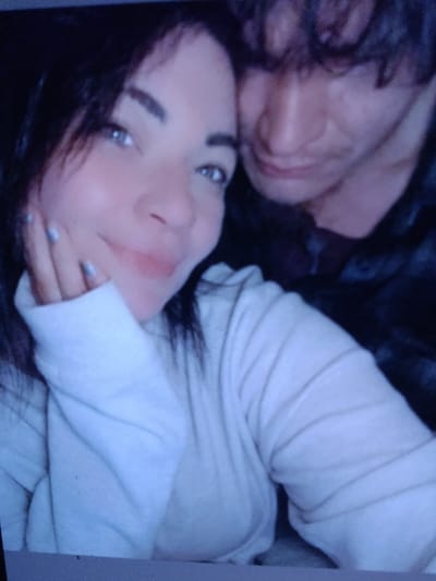 The image displays a close-up photograph of two individuals, appearing to be in an indoor or dimly lit environment. A female subject is prominent in the foreground, facing the camera with a slight smile. She has dark hair and light-colored eyes. She is wearing a light-colored, long-sleeved top. Her right hand is positioned near her face, with light-colored nail polish visible on her fingernails. Behind and to the right of the female, a second individual, appearing to be male, is partially visible. He has dark, curly hair, and his face is partially obscured, with his gaze directed away from the camera. He is wearing a dark-colored garment. The individuals are in close proximity. The background is dark and indistinct, offering no discernible details about the specific environment or location. The precise location within the city of Ozark, United States, is not identifiable from the image.