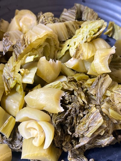 A close-up, overhead view shows a pile of pickled mustard greens in a dark bowl. The greens are a pale yellow-green and appear to be chopped into bite-sized pieces. Some pieces are still attached to stems, while others are separated leaves. The texture of the greens ranges from tender and slightly translucent to dried and wrinkled. The lighting is bright and even, highlighting the details of the food. The bowl appears to be made of metal and has a matte finish. The scene is entirely focused on the food, with no discernible background elements to indicate a specific location. There are no people or animals present in the image.