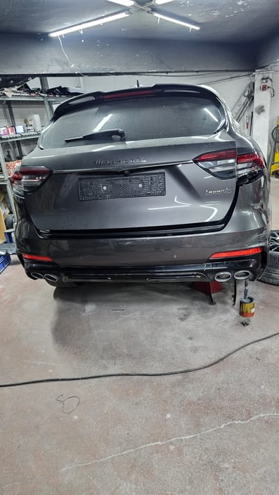 A dark gray Maserati Levante SUV is parked in a garage with its rear facing the camera. The vehicle is raised on a red hydraulic lift. The car's trunk is closed, and the license plate area is visible. The rear bumper has quad exhaust tips and a black diffuser. Red reflectors are present on the lower corners of the bumper. The garage floor is concrete with some visible cracks. In the background, shelves with various items are stacked, and overhead fluorescent lights illuminate the space. A black cable runs across the floor. A small can and some metal parts are on the floor near the exhaust pipes. The location is identified as Ataşehir, Turkey.