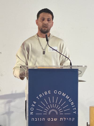 A male individual, appearing to be in his 30s-40s with short dark hair and light facial hair, stands behind a podium. He wears a light beige long-sleeved crew-neck sweater and a silver-colored chain necklace with a rectangular pendant. A white name tag partially visible on his right chest reads "Eliha". His mouth is open, indicating he is speaking. A black gooseneck microphone is positioned in front of him. The podium features a clear acrylic top and a dark blue front panel. The panel displays white text: "NOVA TRIBE COMMUNITY" arched above a stylized sun emblem, and "קהילת שבט הנובה" in Hebrew below the emblem. The background is a plain white wall. The scene is set indoors in Kadima - Zoran, Israel.