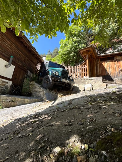 In Shiroka Laka, Bulgaria, an olive-green vintage off-road vehicle, identified by its license plate "CM 7058 MC", is parked on a steep, gravelly incline adjacent to a rustic wooden building. The vehicle is positioned on a stone foundation, with its front tires angled towards the left and its rear tires resting on the ground. The building, constructed of weathered wooden planks, features a prominent pitched roof and an attached wooden shelter. Nearby, a wooden fence encloses an area to the right of the building. A traditional wooden wheel, possibly an antique artifact, lies on the ground to the left of the building's entrance. The scene is bathed in sunlight, casting shadows from the surrounding trees and foliage onto the ground and structures. The sky is clear and blue, with the lush green canopy of trees overhead dappling the scene with light and shade. The ground is a mixture of gravel, dirt, and scattered stones, indicating a rural or semi-rural setting.
