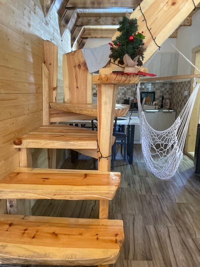 This image depicts an interior scene in Palež, Montenegro, characterized by a rustic and natural wood aesthetic. A prominent, light-toned, natural wood staircase with live-edge steps occupies the foreground and midground, curving upwards from the lower right to the upper left. Black string lights are wrapped around the central support post and banister of the staircase. A small artificial Christmas tree, adorned with red baubles and pinecones, rests on a flat wooden surface extending from the central staircase post, accompanied by a red felt decorative item. A light gray towel is draped over an upper section of the wooden staircase structure. The left wall is clad in horizontal light wood paneling. The floor is composed of variegated brown and gray wood-look tiles. In the background, a kitchen area is visible, featuring a light-colored square-tiled backsplash, white lower cabinets, a sink with a black tray holding items, and a visible cooktop. A white macrame-style hammock chair is suspended from a horizontal wooden rod, hanging over a partially visible white dining table with dark legs and dark chairs. The ceiling incorporates exposed wooden beams, some exhibiting dark, possibly charred, finishes. No individuals are present, and no actions or interactions are depicted.