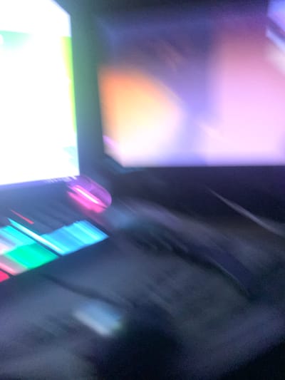 The image shows a blurred view of a computer mouse with pink and purple lights, situated on a keyboard with green and blue lights. The background displays a brightly lit screen with a gradient of orange, purple, and white colors, also heavily blurred. The overall scene suggests a dimly lit environment, possibly a gaming setup or a workspace, with the primary focus on the illuminated computer peripherals. The location in Glodeni, Moldova, is not discernible from the visual information provided in the image.