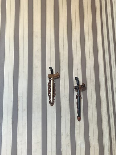 The image displays an indoor wall covered in vertically striped wallpaper, alternating between a light off-white/cream and a medium gray/taupe color. The lighter stripes exhibit a subtle textured or paneled appearance with minor surface imperfections.

Two decorative objects, resembling traditional Indonesian or Malay keris (kris) daggers in scabbards, are mounted vertically on the wall.

The object on the left, positioned slightly left of the image center, features a dark, organically shaped hilt. Its scabbard consists of a light-colored, curved wooden *wrangka* (upper part) and a dark, intricately carved wooden *gandar* (main body) embellished with a central golden-toned metallic strip displaying swirling patterns.

The object on the right, positioned to the right and slightly lower than the left object, is smaller in size. It has a dark, mottled wooden hilt with an organic shape. Its scabbard comprises a mottled light and dark wooden *wrangka* and a slender, dark wooden *gandar* with subtle carved details, tapering to a dark reddish-brown tip.

The objects are stationary, affixed to the wall, serving an ornamental purpose. The lighting is uniform, suggesting an interior setting.