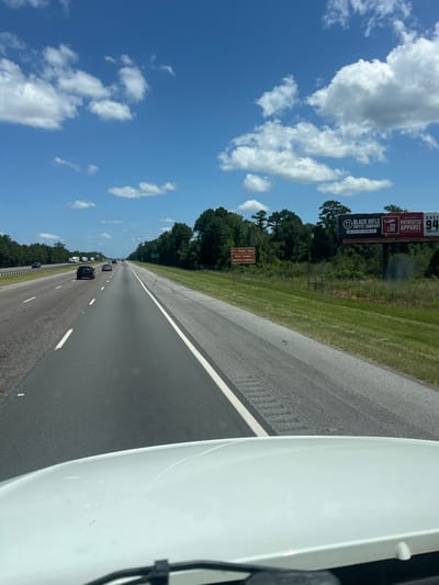 The image captures a view from the perspective of a vehicle traveling on a highway in Riceboro, United States. The road ahead is asphalt, with multiple lanes and white lane markings. Several vehicles are visible on the road, including cars and trucks. The environment features a bright blue sky with scattered white clouds. On the right side of the highway, a grassy median is present, followed by a fence and a dense line of trees. A brown sign is visible on the left side of the median, reading "R.V. 'Bobby' Sikes Fallen Troopers Memorial Highway." Further down the road on the right, a large billboard displays advertisements for "Black Rifle Coffee Company" and "Nine Line Patriotic Apparel." The foreground shows the white hood of the vehicle from which the image is taken, with a portion of the windshield wipers visible. The lighting suggests it is daytime, with strong sunlight casting shadows.