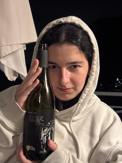 An individual is depicted holding a bottle of wine, captured in a medium close-up at night in Nesso, Italy. The subject is a young adult female with dark hair, wearing a light-colored hooded sweatshirt over a black collared top. She holds a dark green glass wine bottle with both hands, positioning it vertically such that the bottle's neck and upper body partially obscure her left eye and forehead. The bottle features a black label with white text, including "pietrErose" and "CANTINE ANGELINETTA Lago di Como", along with a stylized white drawing of two figures and a grape leaf logo. The subject looks directly at the camera with a neutral expression. In the background, a large white fabric structure, likely an awning or umbrella, is visible on the upper left. The distant background is dark, indicating nighttime, with faint light sources suggesting a view over a landscape.