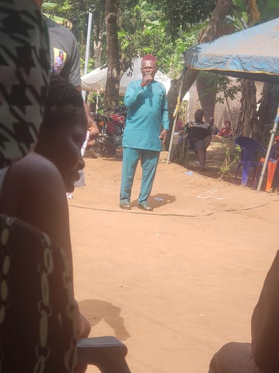 This outdoor image captures a community gathering or event under bright daylight in what appears to be a sunny, rural or semi-rural setting, consistent with Umundugba, Nigeria.
The central figure is a man standing on unpaved light brown earth, actively speaking into a microphone. He is dressed in a traditional long-sleeved teal tunic with matching trousers and a maroon cap. His attire suggests a formal or community leadership role.
An audience is present, with several individuals visible in the foreground and background. In the immediate foreground, the back of a person's head and shoulder are seen, adorned with braided hair and wearing a dark, possibly patterned, garment. Another person's leg and foot are partially visible in the extreme lower right.
In the background, on the right side, several people are seated on blue and orange plastic chairs. One person behind them wears a black t-shirt with an indecipherable graphic print. Further back, a red brick wall is visible. To the left and behind the speaker, a red motorcycle is parked next to a tree. Lush green trees and foliage frame the left side of the scene.
A light blue tarp or canopy stretches across the upper left portion of the image, providing shade. The strong shadows cast by the subjects and objects indicate a bright, sunny day. The event appears to be a public address or a community meeting, with the speaker engaging an assembled audience. No discernible text is clearly visible on clothing or objects, other than a pattern on the speaker's shirt and an unreadable graphic on a background figure's shirt.