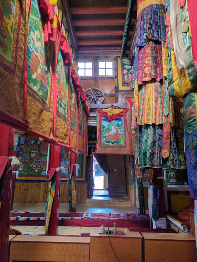 This image shows the interior of a room within a building, likely a monastery, in Yulang, India. The room is narrow and long, with high ceilings supported by wooden beams. The walls are adorned with colorful thangka paintings and intricate fabric hangings, predominantly in red, yellow, blue, and green hues, featuring floral and religious motifs. Red wooden pillars line the sides of the room, supporting horizontal beams from which more fabric decorations hang. In the distance, a doorway leads to another section of the building, and two square windows are visible high on the wall, allowing natural light to enter. The floor is made of wooden planks, and red mats are laid out, suggesting a space for meditation or prayer. In the foreground, low wooden tables are present, and on one of these tables, a microphone and some bells are visible, indicating the presence of sound equipment and possibly a ceremonial setup. The overall atmosphere is serene and rich with religious and cultural artifacts. There are no people visible in the image.