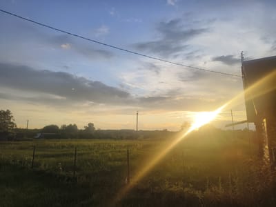 The image depicts a rural landscape at sunset, likely in the vicinity of Кругліцкі сельскі Савет, Belarus. The sky is a mix of blue and grey clouds, with the sun setting on the right side of the frame, casting a strong golden ray across the scene. In the foreground, a grassy field stretches out, with some sparse vegetation and a faint fence line. A wooden structure, possibly a building or shed, is visible on the far right, partially silhouetted against the bright sunset. A utility pole stands in the middle ground, and a line of trees forms the horizon. The overall impression is one of a peaceful, late-day rural setting.