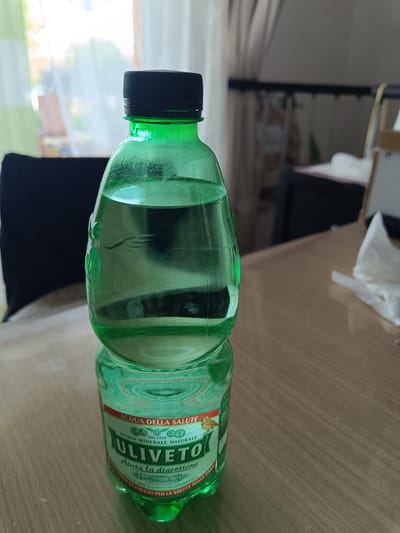 This is a close-up, eye-level shot of a green plastic bottle of Uliveto mineral water sitting on a wooden table. The bottle has a black cap and is filled with clear liquid. The label on the bottle is predominantly white and green, with text in Italian. Visible text includes "ACQUA DELLA SALUTE", "ULIVETO", "ACQUA MINERALE NATURALE", "Aiuta la digestione", and "FONTE DI CALCIO PER LA SALUTE DELLE OSSA". A small illustration of a bird is also visible on the label.

The background is softly blurred, showing an indoor setting. Through a window with vertical blinds, daylight is entering, suggesting it is daytime. The blinds appear to be light green and white. To the left of the bottle, a dark object, possibly a chair or part of furniture, is visible. To the right, a white paper napkin is placed on the table. The overall lighting suggests a moderately bright environment, possibly a home or restaurant. The scene is static, with no people or animals actively engaged in any activity. The focus is clearly on the water bottle. The wooden table surface has a visible grain pattern and a warm, light brown tone. The reflection of the bottle is visible on the table.