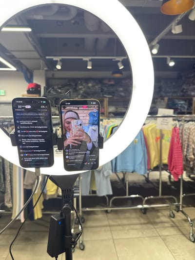 The image captures a close-up of a live-streaming setup, likely in a retail store environment. A large, bright white LED ring light is prominent in the foreground, with two smartphones mounted vertically within its circumference.

The top smartphone displays a live video feed of a young woman. She has light-colored hair and is wearing a beige or light-colored headband or head covering. She holds another smartphone with a distinctive floral phone case, appearing to be taking a selfie or recording herself. She is smiling, and her manicured nails are visible. The background of her video shows what looks like a wooden surface or floor, and possibly some hanging denim clothing. The top left corner of her screen shows the time "17:29" and the word "LIVE" in red.

The bottom smartphone displays a live chat or comment section, presumably for the stream shown on the top phone. It also shows the time "17:29" in the top left corner. The chat feed contains multiple entries, predominantly in German, indicating viewers joining the stream and commenting. Visible messages include:
*   "petraschutz ist beigetreten" (petraschutz has joined)
*   "petraschulz erneut veröffentlicht" (petraschulz reposted/published again)
*   "Goldmarie 1408 ist beigetreten" (Goldmarie 1408 has joined)
*   "Leiterlily ist beigetreten" (Leiterlily has joined)
*   "Manusia ist beigetreten" (Manusia has joined)
*   "Mehr Likes können Zuscha" (More likes can [audience/viewer]...)
*   "ver innen anziehen. Weiter" (attract inside. Further...)
*   "sol"
*   "Lisa ist beigetreten" (Lisa has joined)
*   "itsme_jisav ist beigetreten" (itsme_jisav has joined)
*   "Anja Neit ist beigetreten" (Anja Neit has joined)
*   "Bella hat dem LIVE einen Like gegeben" (Bella liked the LIVE)
A "GR" logo or text is visible in the top right corner of this phone's screen.

Both phones are held in place by a black mounting bracket attached to the ring light's stand. Cables are visible, with one extending from the bottom phone and another connected to a black power bank or adapter positioned on the right side of the stand.

In the background, out of focus, are clothing racks holding various garments in colors such as pink, yellow, light blue, and light green, suggesting the setting is a clothing store or boutique. Overhead lights are reflected in the shiny surface of the ring light and potentially a mirror. The floor appears to be made of light-colored tiles. The overall impression is that of a professional or semi-professional setup for a social media live stream, possibly for fashion or product promotion.
