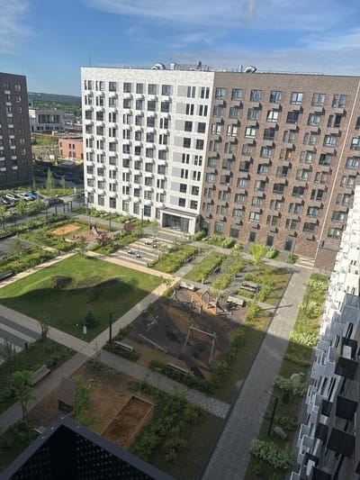 This is an aerial, slightly angled, medium-shot photograph of a modern residential courtyard in the city of Urozhay Spb, Russia. The scene is dominated by two multi-story apartment buildings, one with a white facade and the other with a brown brick facade, flanking a communal courtyard. The courtyard features a playground with swings and a slide, a sandbox, and a small wooden playhouse. Paved walkways crisscross the courtyard, separating grassy areas and landscaped garden beds with small trees and bushes. Benches are strategically placed throughout the courtyard for seating. In the background, other buildings and some trees are visible. The sky is blue with scattered clouds. The overall impression is one of a well-maintained, contemporary urban living space.