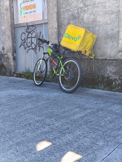 A Glovo delivery bicycle leans against a concrete wall next to a boarded-up building in Cangas de Morrazo, Spain. The bicycle is green with white rims and has a large, yellow Glovo delivery bag attached to the rear rack. The wall is weathered and covered in moss. The boarded-up building has a "VENDE" (For Sale) sign and graffiti on the corrugated metal. The ground is concrete with sunlight reflecting on it.