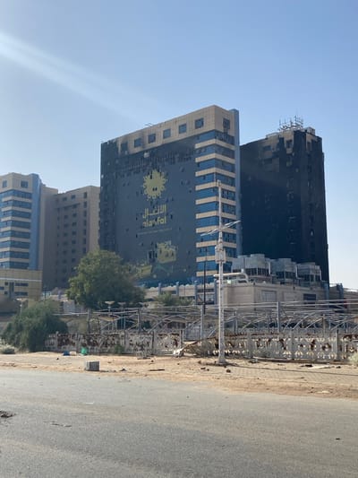 The scene is a street in Khartoum, Sudan (latitude: 15.602, longitude: 32.525).  The street is empty and paved with asphalt.  A fence made of metal bars runs along the edge of the street, and some of the bars are broken and bent.  There is rubble on the ground in front of the fence, and some green foliage is growing in the dirt.  Behind the fence, there are three large buildings.  The building in the center is the tallest and appears to be a commercial building with an advertisement on the side featuring a large yellow sun with an Arabic logo reading "Al-Asfal" in yellow letters. The building is heavily damaged, with many windows broken and missing.  The building to the left of the center building is also heavily damaged, with many of its windows broken and missing.  The building to the right of the center building is also damaged, with many of its windows broken and missing.  The buildings are all painted a dark gray color, and they appear to have been the target of a bombing or other type of violent attack.  There is a white metal pole in the center of the scene with a white metal light fixture on top of it. There is no evidence of people or vehicles in the scene.