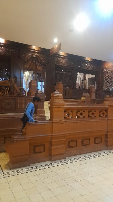 This image captures the interior of a traditional Indonesian building, consistent with architectural styles found in areas such as Glantengan, Indonesia. The scene features a lone individual, appearing male, in the left midground. They are wearing a light blue long-sleeved shirt and dark trousers, with a black shoulder bag, and are observed leaning forward with their left hand resting on a light-colored wooden balustrade, seemingly in the process of stepping onto or over it.
The environment is characterized by richly carved dark wood structures that form walls, doorways, and decorative panels in the background, displaying intricate traditional patterns. To the far left, a large, detailed wooden sculpture resembling a vintage motorcycle is visible. The foreground features a light-colored, ornate wooden balustrade with geometric cutouts. The floor is composed of light-colored, possibly beige or off-white, square tiles with a dark, patterned border. The ceiling is white and appears high, illuminated by multiple recessed lights, with one light source in the upper right casting a prominent glare. The overall impression is that of a meticulously decorated museum, exhibition space, or a grand traditional hall.