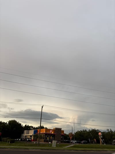 An exterior, eye-level, medium shot captures a Dairy Queen restaurant in Bradenton, Florida, under a cloudy, overcast sky. The restaurant, with its signature orange and blue facade, is situated on the right side of the frame. A line of cars is visible in the drive-thru. Several power lines traverse the sky horizontally across the image. In the background, a dense line of trees forms a dark silhouette against the lighter sky, with a patch of lighter, potentially rain-filled clouds forming in the distance. A utility pole stands in front of the Dairy Queen. The foreground shows a grassy median and a road.