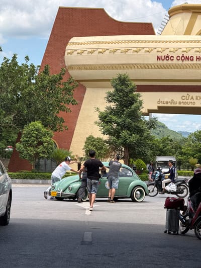 In front of a building with red brick and beige accents, a group of people are gathered around a vintage green Volkswagen Beetle. The car is being pushed by three individuals. The building has a large archway with decorative elements and text in Vietnamese. To the right, two men are seated on motorcycles. One motorcycle is being ridden by a man holding a bag, and another motorcycle is parked nearby. The scene takes place on an asphalt road with some foliage and trees visible in the background, suggesting an outdoor urban or border crossing location. The overall impression is of a functional setting, possibly a border checkpoint, given the presence of vehicles and the official-looking building.