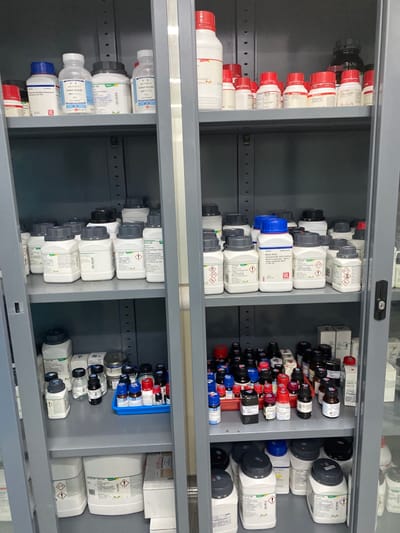 The image displays the interior of a tall, gray metal storage cabinet, likely situated within a laboratory or chemical storage facility in Cipeundeuy, Indonesia. The cabinet features four shelves, each densely stocked with various chemical reagents and containers. The two central doors of the cabinet appear to be glass, providing visibility of the contents.
On the top shelf, a collection of white plastic bottles with blue, white, black, and red screw caps are arranged. Several labels with product names such as "Sodium Acetate" and "Iodide Acetate" are discernible. The second shelf from the top is filled with numerous larger white plastic containers, predominantly with gray or black screw caps, many displaying green-bordered labels with an "M" logo and GHS hazard pictograms, including an exclamation mark (GHS07) and flame over circle (GHS03). Some bottles on this shelf have blue caps. The third shelf from the top contains a multitude of smaller bottles, some made of clear glass, others of dark plastic, with various colored caps (red, blue, black). Many of these smaller bottles are organized within blue and red plastic trays. The bottom shelf holds more large white plastic containers, similar to those on the second shelf, featuring gray, black, or yellow screw caps, "M" logos, and hazard pictograms. No individuals are present, and no actions or interactions are depicted within the static scene.