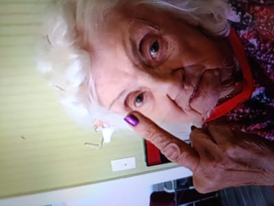 The image is a close-up, tilted shot of an elderly woman's face and hand. She has white hair and is wearing a patterned shirt with a red collar. Her right index finger is raised, with a vibrant purple nail polish. The background appears to be a room with pale green walls and some objects on a surface, including what looks like a light switch and a small white card. The lighting suggests it might be daytime indoors. The woman's expression is difficult to discern clearly due to the tilt and resolution, but her eyes are open. The overall impression is informal, possibly a selfie or a candid photo taken by someone close to her. There is no visible text in the image. The location context provided is Krasivka, Ukraine.