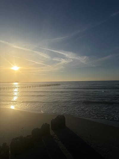 The image depicts a beach scene in Ustka, Poland, during a sunset. The sun is low on the horizon, casting a golden glow across the water and sand. The sky is a gradient of blue and yellow, with wispy clouds. The sea is calm, with small waves lapping at the shore. In the foreground, the dark silhouettes of wooden breakwaters emerge from the sand. In the middle ground, a line of similar breakwaters extends into the sea. A distant structure, possibly a lighthouse, is visible on the coastline. The overall mood is serene and peaceful, capturing the tranquil beauty of a coastal sunset.