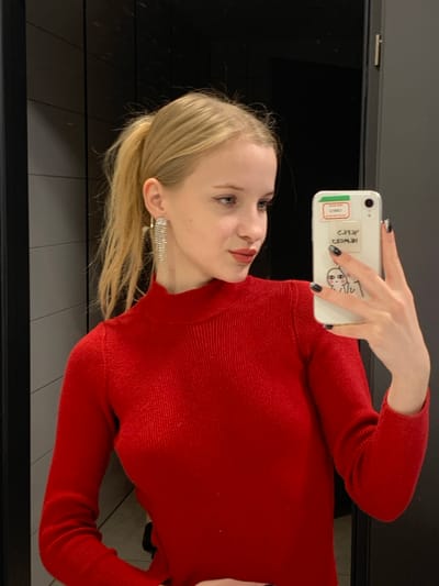 A young female subject with light skin and blonde hair styled in a high ponytail is captured in a mirror selfie. She is wearing a red, ribbed, long-sleeved turtleneck top. Her left ear is adorned with a long, multi-strand, sparkling earring. Visible makeup includes red lipstick and winged eyeliner. The subject's right hand holds a white smartphone, likely an iPhone, which has a clear or white case featuring several stickers: "CTM007", mirrored text "393H 2MOWOH", and a graphic of two simplified faces. The visible fingernails on her right hand are painted black. The background consists of dark, possibly black or very dark grey, paneled surfaces with horizontal and vertical seams, indicating an indoor setting. The scene is reportedly located in Nowy Targ, Poland.