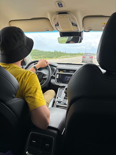 The image shows the interior of a car from the perspective of a rear passenger. A person wearing a yellow t-shirt and a black bucket hat is driving. The car is on a highway with trees and greenery on either side. The sky is overcast. The car's dashboard and infotainment screen are visible, showing the song "COLD WAVES" playing. A black SUV is visible in front of the car, also on the highway. This scene is consistent with driving on a highway in a suburban or rural area, and could be near Красная Горка, Russia, given the presence of similar landscapes in that region.