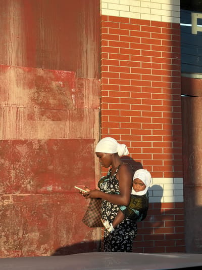 An over-the-shoulder shot captures a Black woman wearing a white headscarf and a black floral dress, standing against a backdrop of a weathered red wall and a red brick building in Muxara, Mozambique. She holds a smartphone in her right hand, looking down at it. A baby, also wearing a white headscarf, is strapped to her back in a colorful wrap, its face turned towards the viewer. The sunlight casts a warm glow on the scene, highlighting the textures of the walls and the subjects. The lower part of the image is obscured by the edge of a car.