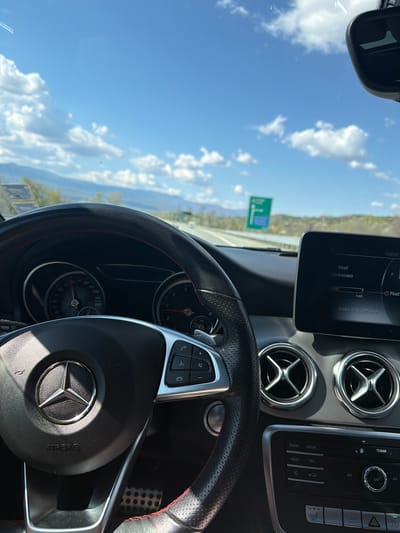 The image shows the interior of a Mercedes-Benz car, focusing on the dashboard, steering wheel, and a view through the windshield. The sky is blue with some clouds. The car's infotainment screen displays music information, and the dashboard features air vents. A road sign is visible through the windshield. The location in Valkovo, Bulgaria is not discernible from this image.