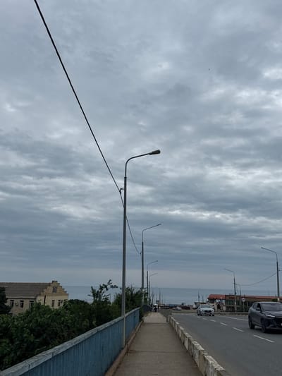 This media file depicts a cloudy day in Derbent, Russia. The scene is an outdoor urban environment with a street, sidewalk, and a body of water in the distance. A power line runs diagonally across the frame. Several streetlights are visible, suggesting it is either dawn, dusk, or an overcast day. Cars are present on the road, indicating typical urban activity. The architecture of a building in the background appears to be consistent with Russian style. The overall mood is neutral, dominated by the overcast sky and mundane street elements. There are no people prominently visible, and the focus is on the urban landscape and the atmospheric conditions. The time of day is not clearly defined but is likely during daylight hours due to the activity on the street. The weather is clearly cloudy.