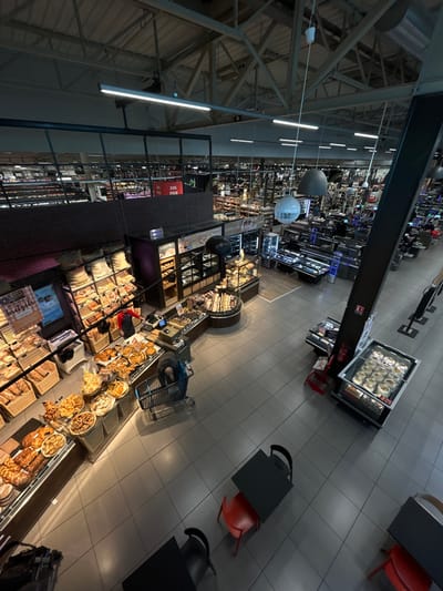 The image presents an overhead view of a large supermarket or hypermarket interior, identified as being located in Eschau, France. The scene captures a wide retail space characterized by high ceilings with exposed structural elements and industrial lighting.
In the foreground, a light gray tiled floor leads to two small seating arrangements, each consisting of a black-topped table and a single chair (one red, one black). To the right, two vertical refrigerated display cases contain various packaged food items. A thick black structural column stands centrally on the right, bearing a red fire extinguisher.
The midground is dominated by an extensive bakery section. A male individual, dressed in a red shirt and dark apron, stands behind a long counter showcasing numerous baskets and trays of breads and pastries. To the left of this counter, a customer wearing dark clothing is bent over a shopping cart, actively selecting items from a display of baked goods. Adjacent to the counter are curved glass display cases filled with smaller pastries, cakes, and tarts. Signs visible in this area include "Le pain bon" and advertisements for "BRETZELS." Stacked bags, possibly of flour, are visible on shelves behind the main bakery display.
Further into the background, multiple retail aisles extend, stocked with various merchandise. Several operational checkout lanes are discernible, with customers and staff present. The ceiling above features an array of exposed metal trusses, beams, and linear fluorescent light fixtures, complemented by large spherical pendant lamps. A sign displaying "DES PRIX" (Prices) is visible above a brick-textured partition wall in the upper left midground, indicating a promotional area.