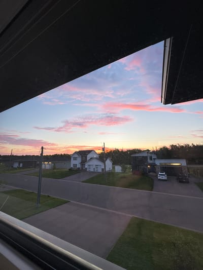 The image depicts a sunset view over a suburban street in Dieppe, Canada. The sky is filled with streaks of pink and orange clouds against a gradient of blue. In the foreground, a dark, horizontal structure, possibly a window frame or awning, partially obscures the view. The street is paved, with manicured lawns on either side. Several houses are visible, characterized by modern architecture with white and dark exteriors, and garages. Two vehicles, a white SUV and a dark sedan, are parked in the driveway of one of the houses. Utility poles and wires are present along the street. The overall scene suggests a peaceful evening in a residential area.
