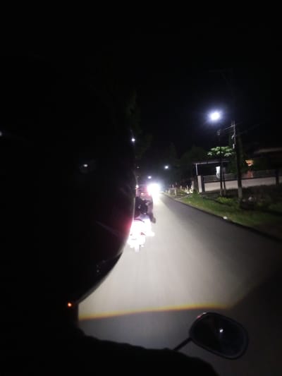 A first-person perspective shot taken at night shows a person riding a motorcycle on a paved road in Walennae, Indonesia. The rider's helmet is prominently featured in the foreground, obscuring a significant portion of the left side of the image. The road ahead is illuminated by the headlights of the rider's motorcycle and other vehicles. Several other motorcycles are visible on the road, some with their headlights on. The surrounding environment includes trees and houses lining the street. A streetlight can be seen in the distance. The overall atmosphere is dark, typical of a nighttime scene in a residential area.