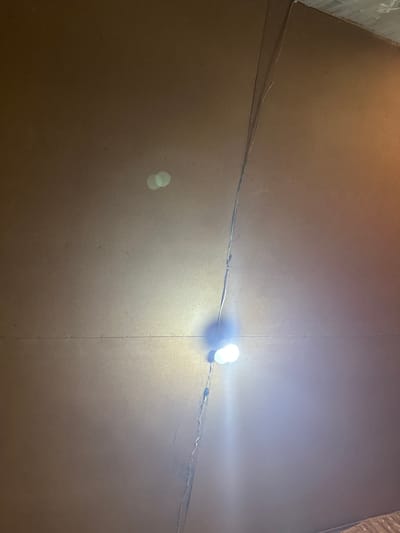 A close-up, overhead shot captures a textured, brown surface, possibly a ceiling, in what appears to be an interior space. A prominent, vertical seam or crack runs down the center of the frame, with a thin electrical wire following its path. Two bright, white LED bulbs are illuminated, casting a strong light on the surface and creating a visible lens flare. The overall lighting is dim, with warmer tones appearing on the right side of the image, suggesting an artificial light source. The surface shows some minor imperfections and dust particles. No people or clear geographic indicators are visible.