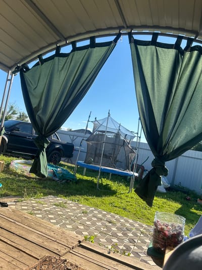 The image depicts a backyard scene on a sunny day, likely in the summer. The foreground is dominated by a green canopy, possibly part of a gazebo or patio cover, with its fabric tied back. Beyond the canopy, a trampoline with a safety net is visible in the middle ground, set up on a green lawn. A dark-colored car is parked behind the trampoline.
In the background, a white fence encloses the yard, and some trees can be seen. To the right of the image, there is a paved area with patterned tiles and wooden decking. A small rug with an oriental pattern is placed on the wooden deck. A clear plastic container filled with red berries, possibly strawberries, is on the paved area.
The overall impression is one of a casual, outdoor space used for recreation and relaxation. The bright sunlight suggests it is daytime, likely mid-morning or afternoon. The presence of a trampoline and a small inflatable pool hints at activities suitable for warm weather. The location is identified as Lotoshino, Russia. There is no visible text in the image.