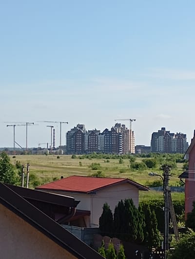 The image depicts a suburban or developing area with a clear blue sky. In the mid-ground, several tall tower cranes are visible against a backdrop of a developing construction site featuring multi-story buildings with varied architectural styles and colors, including brick and white facades. Some of these buildings appear to be apartment complexes or residential blocks. Further back, more construction cranes and industrial-looking structures are discernible. The foreground shows residential rooftops, one with a red tile roof and another with a dark brown angled roof, along with evergreen trees and a utility pole with a street light. The environment appears to be a mix of ongoing construction and established residential areas. The scene is set in Maloye Isakovo, Russia, as indicated by the request, likely a semi-rural or exurban locale experiencing development.