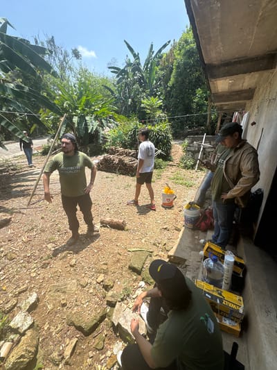The scene shows a group of people engaged in an outdoor activity in Pinahuista, Mexico. The location is a partially shaded area under some sort of overhang, possibly a building structure.

Four people are clearly visible. One man wearing a green t-shirt stands in the foreground, gesturing towards the ground. He's holding a stick. Another person in a white shirt and tan shorts is standing behind the man. Near the overhang, a man wearing a brown jacket is visible. Adjacent to the man in the brown jacket, a person wearing a black hat and a green t-shirt appears to be working on something on the ground.

Objects scattered around the area include DeWalt boxes, a plastic bottle, a bucket filled with yellow material, and a red bag.

The environment consists of a gravelly ground, vegetation, including banana trees, and a bright blue sky.