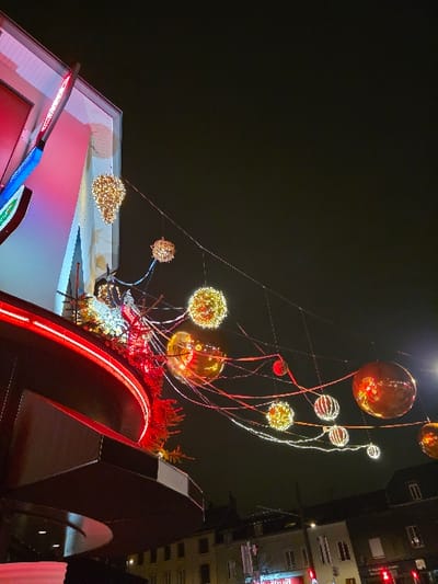 Nighttime view of festive decorations. Illuminated spheres of varying sizes ascend on thin cables. Below, a building roof is visible, adorned with red lighting and decorative reindeer figures. A prominent architectural structure with a red illuminated arch dominates the foreground, juxtaposed against taller buildings in the background.