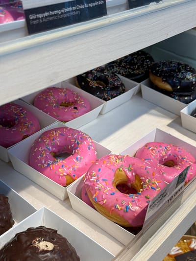 This image shows a display of donuts in what appears to be a bakery. The donuts are arranged in white paper trays on wooden shelves. There are pink iced donuts with colorful sprinkles, and dark chocolate iced donuts with sprinkles. In the background, there is a sign with Vietnamese text that reads "Giữ nguyên hương vị bánh mì Châu Âu nguyên bản" and "Fresh & Authentic European Bakery." There is also a price tag visible on one of the pink donuts. The overall environment suggests a retail setting, likely a donut shop or bakery, in Phu Thuy, Vietnam. No people are visible in the image.